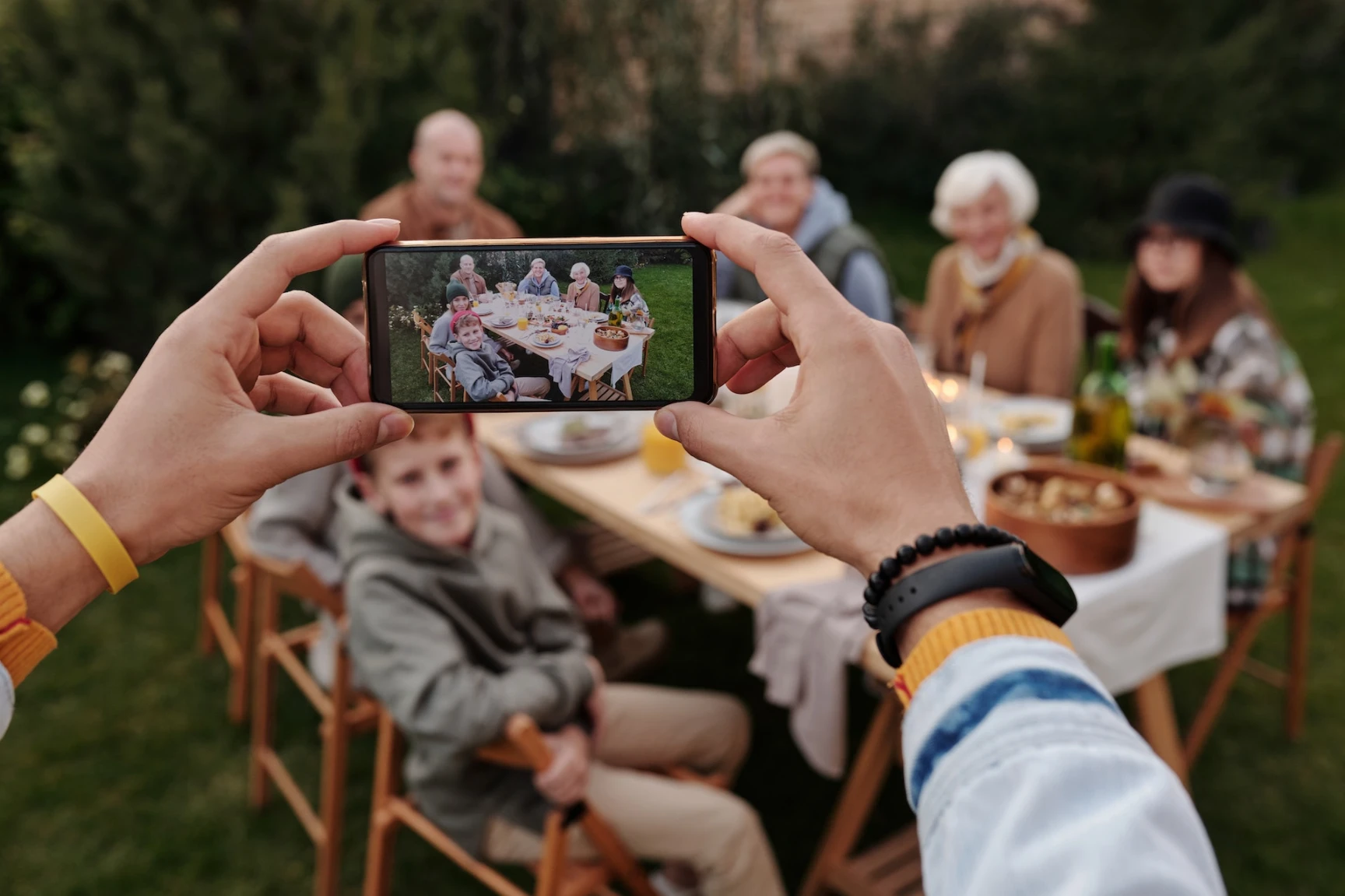 multiple generations of family members slightly out of focus sitting at an outdoor dining table, one person in the foreground has them composed for a photo on their cellphone